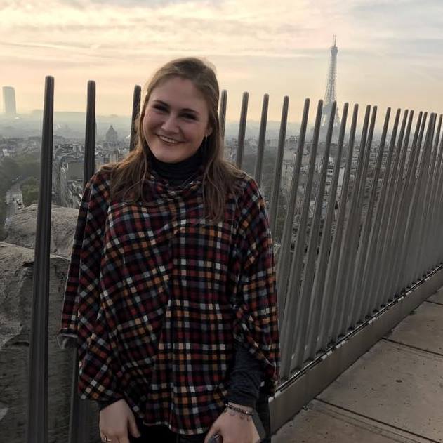 Lauren Fick is smiling in front of a fence in front of the Eiffel Tower. She is wearing a red check peacoat and black turtleneck.