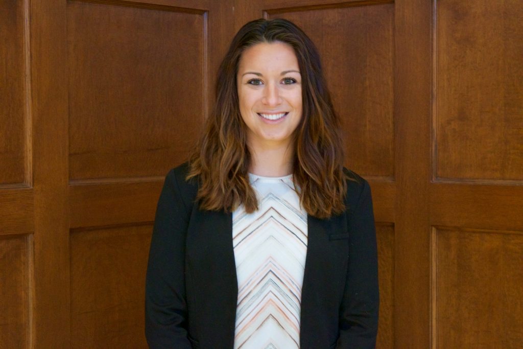 Elizabeth Picca is smiling in front of wooden panels. She is wearing a dark blazer with a blouse with an upward-facing Chevron pattern.