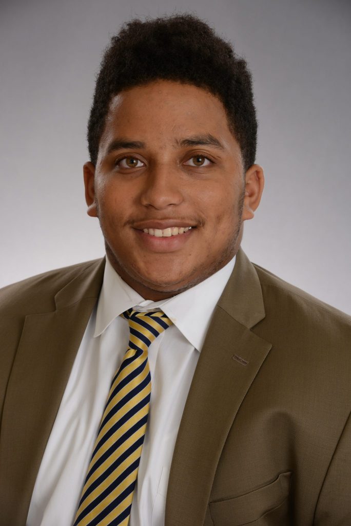 Steven Bradley smiles in front of a grey background. He has dark hair, and is wearing a light brown suit with a yellow and black striped tie.