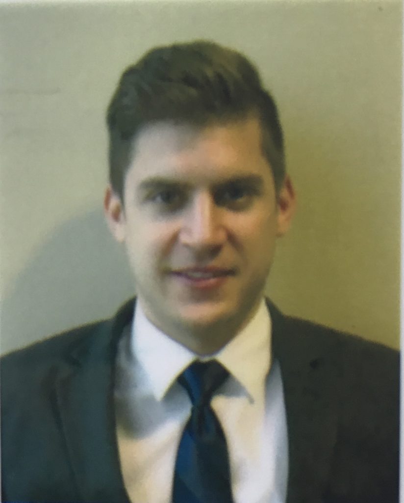 Eleftherios Lazaridis is standing in front of a wall in a dark suit and black tie.