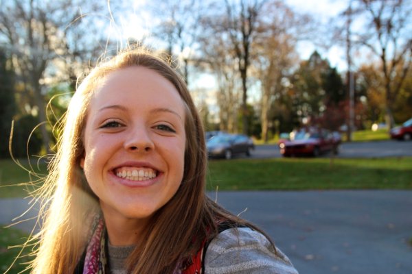 Grace Davis is smiling in front of a parking lot and trees with the sun shining on her cheek.