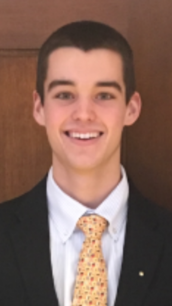 George Litman is smiling in front of wooden panels. He has dark hair, and is wearing a dark suit with a yellow, patterned tie.