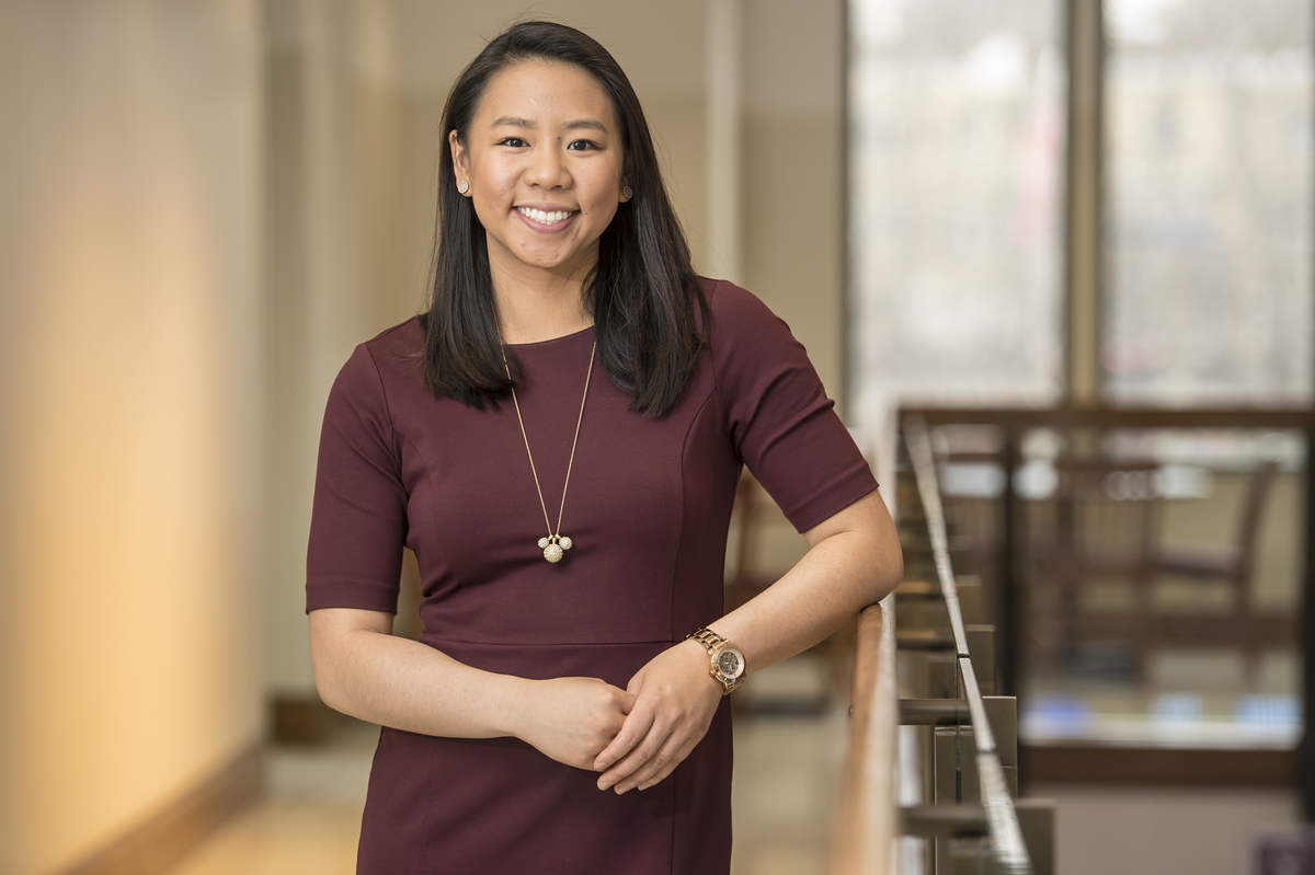 Morgan Bui is smiling in a blurred hallway, leaning against a railing. She is wearing a dark red dress with a gold necklace.