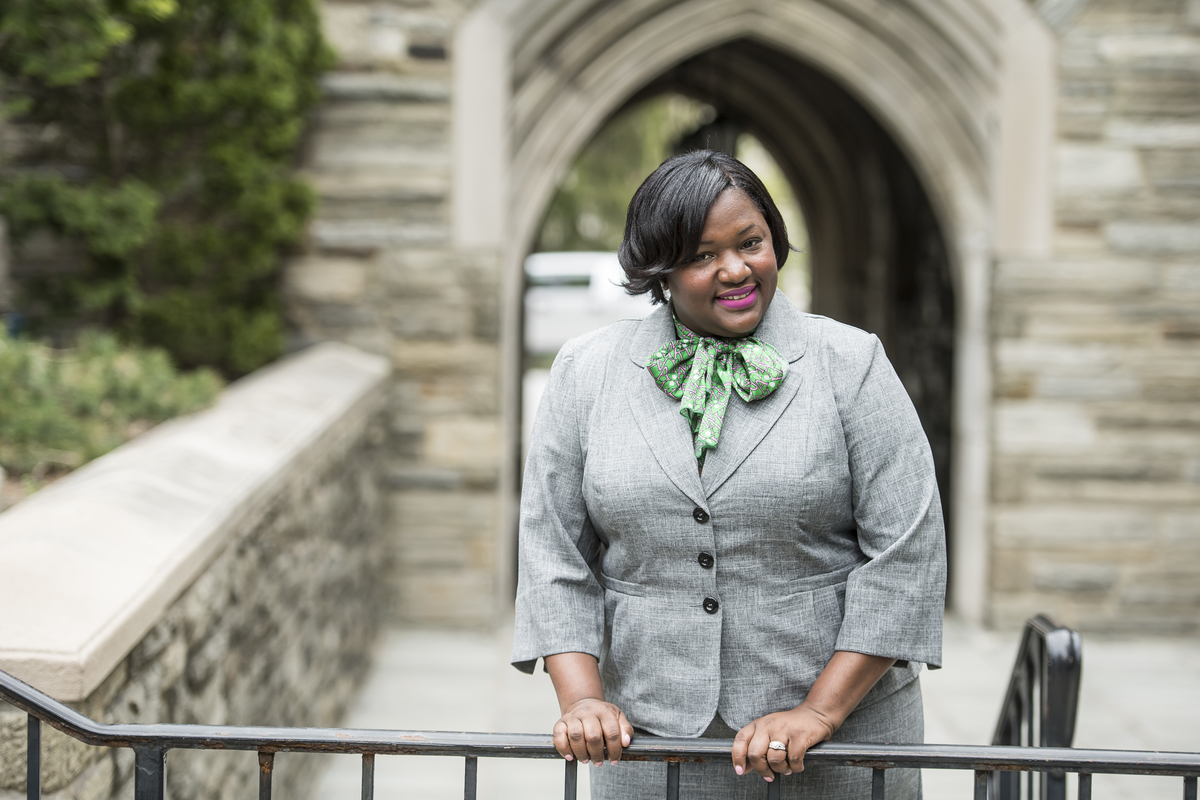 Nyshawana Francis-Thompson is smiling in the Barbelin courtyard. She is wearing her hair short, with a light grey suit and pale green scarf.