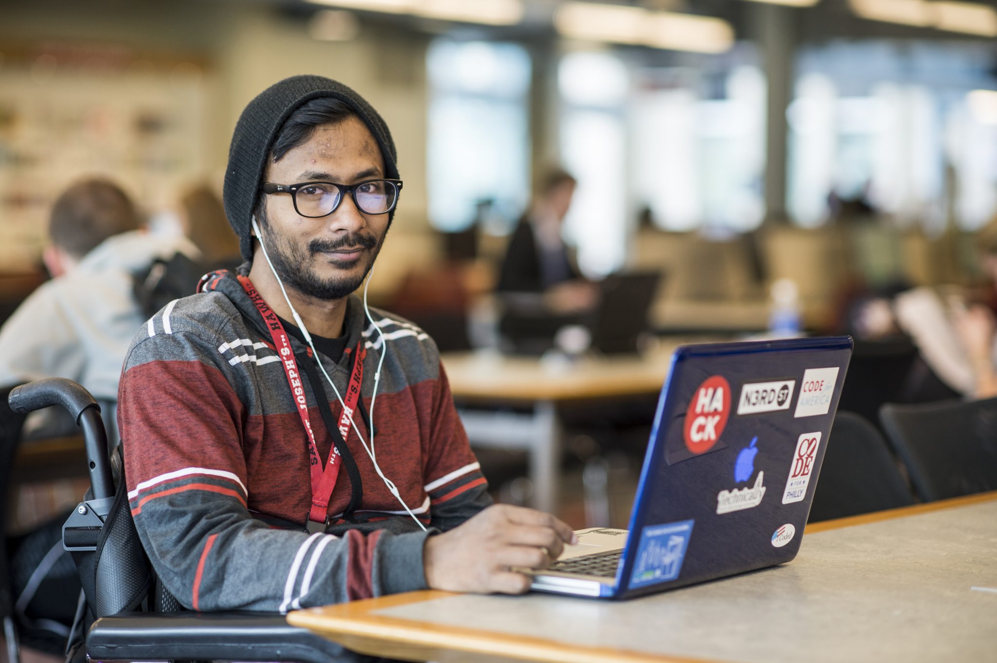 Ather Sharif is sitting in the library in front of his blue laptop covered in stickers. He is wearing a black beanie, striped hoodie, a lanyard, and has white earphones in his ears.