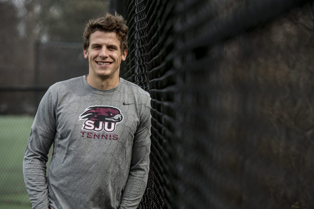 Kyle Chalmers is leaning against a chain link fence, wearing a grey SJU Tennis long-sleeved shirt.