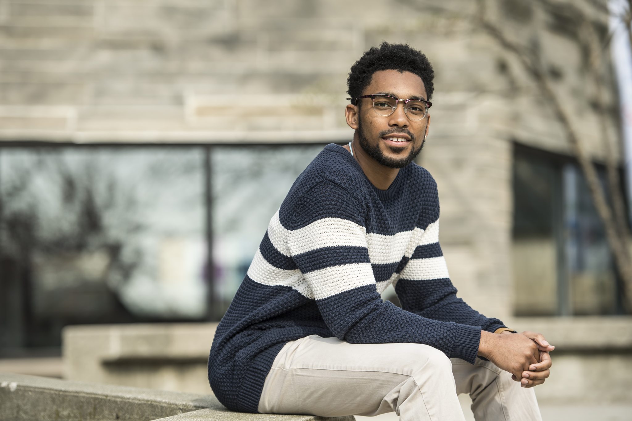 Prince Yakubu is sitting outside in front of a stone building and bare trees. He is wearing a grey shirt with two white stripes, glasses, and has light slacks.