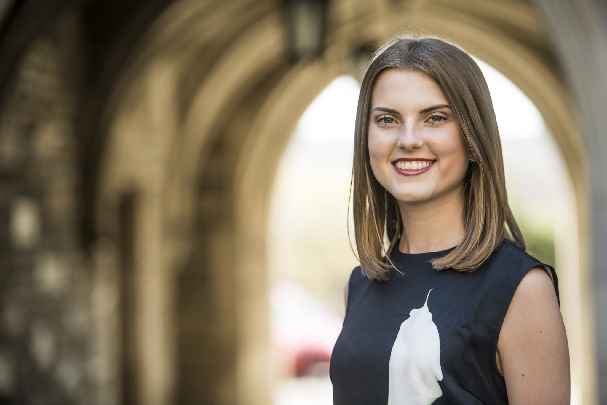 Aly Homan is standing in the blurred archway of Barbelin. Her hair is straight, and she is wearing red lipstick and a black and white blouse.