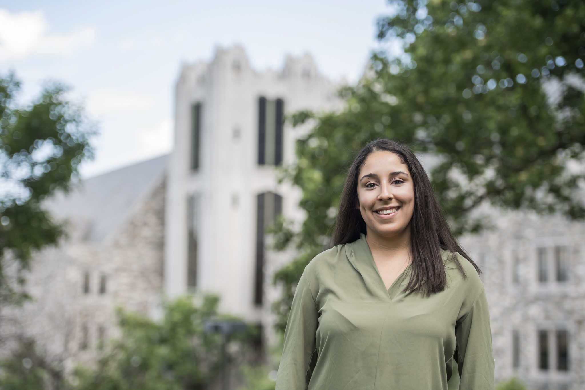 Elizabeth Fuentes is smiling in front of a blurred Mandeville Hall. She is wearing a light green top with her hair straight.