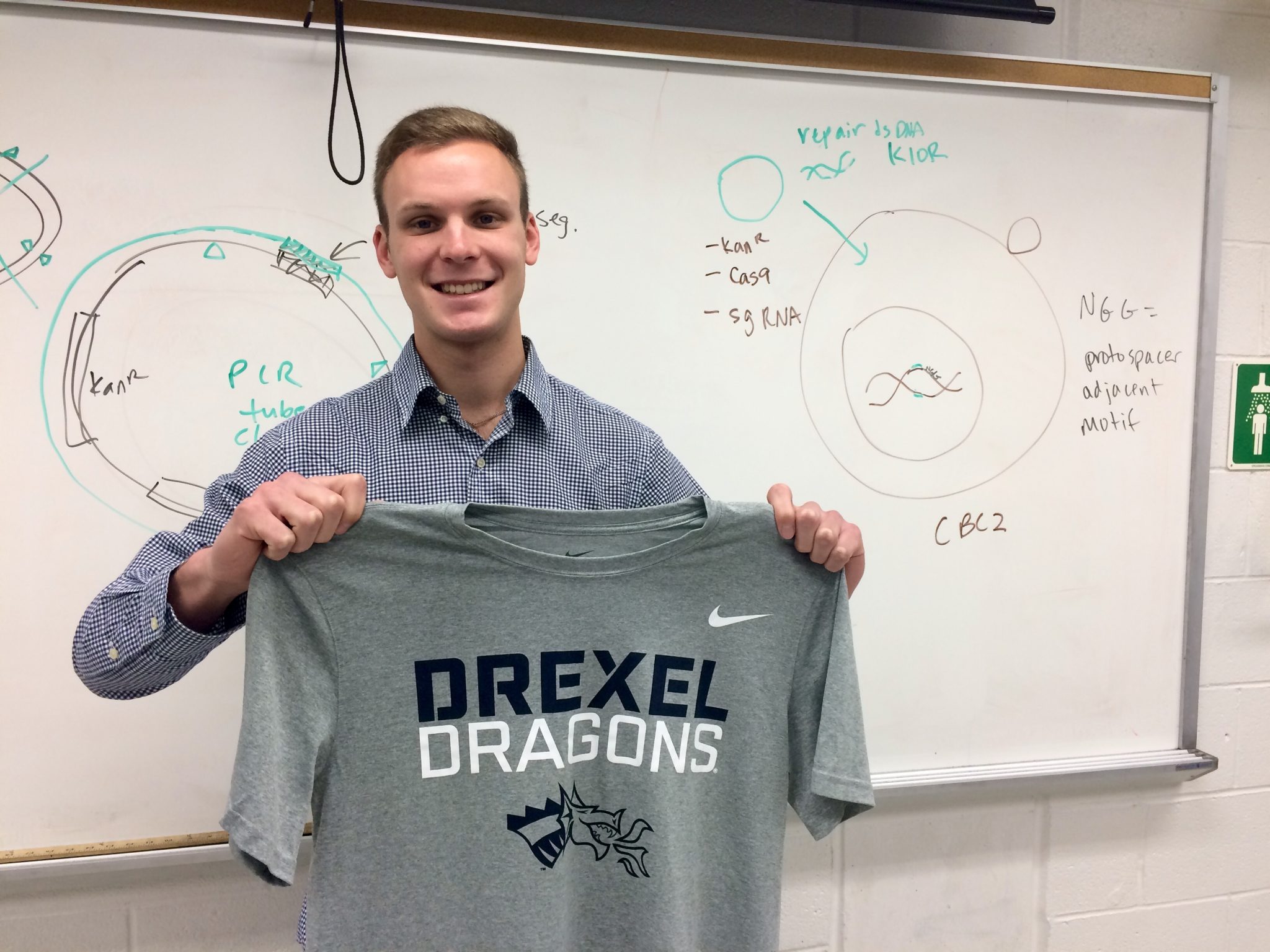 Andrew Arner is smiling in front of a whiteboard with notes and diagrams. He is holding a grey shirt that reads, "Drexel Dragons."