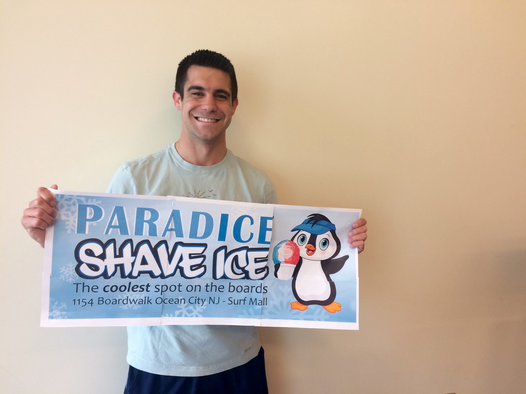 Bobby Lattanzi is smiling and holding a sign with a small penguin with a visor that reads, "Paradice Shave Ice."