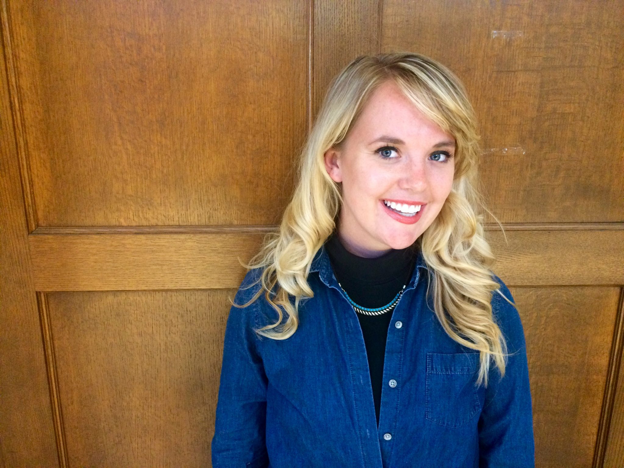 Clare Kocman is smiling in front of wooden panels. She is wearing a black turtlenecks with a blue top, and a teal and gold necklace.