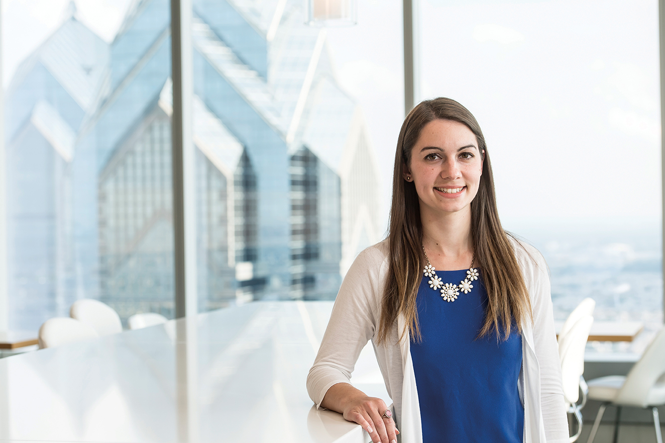 Rebecca Rosati is smiling in front of a window overlooking various glass office buildings. She is wearing a white cardigan, blue top, and white floral necklace.