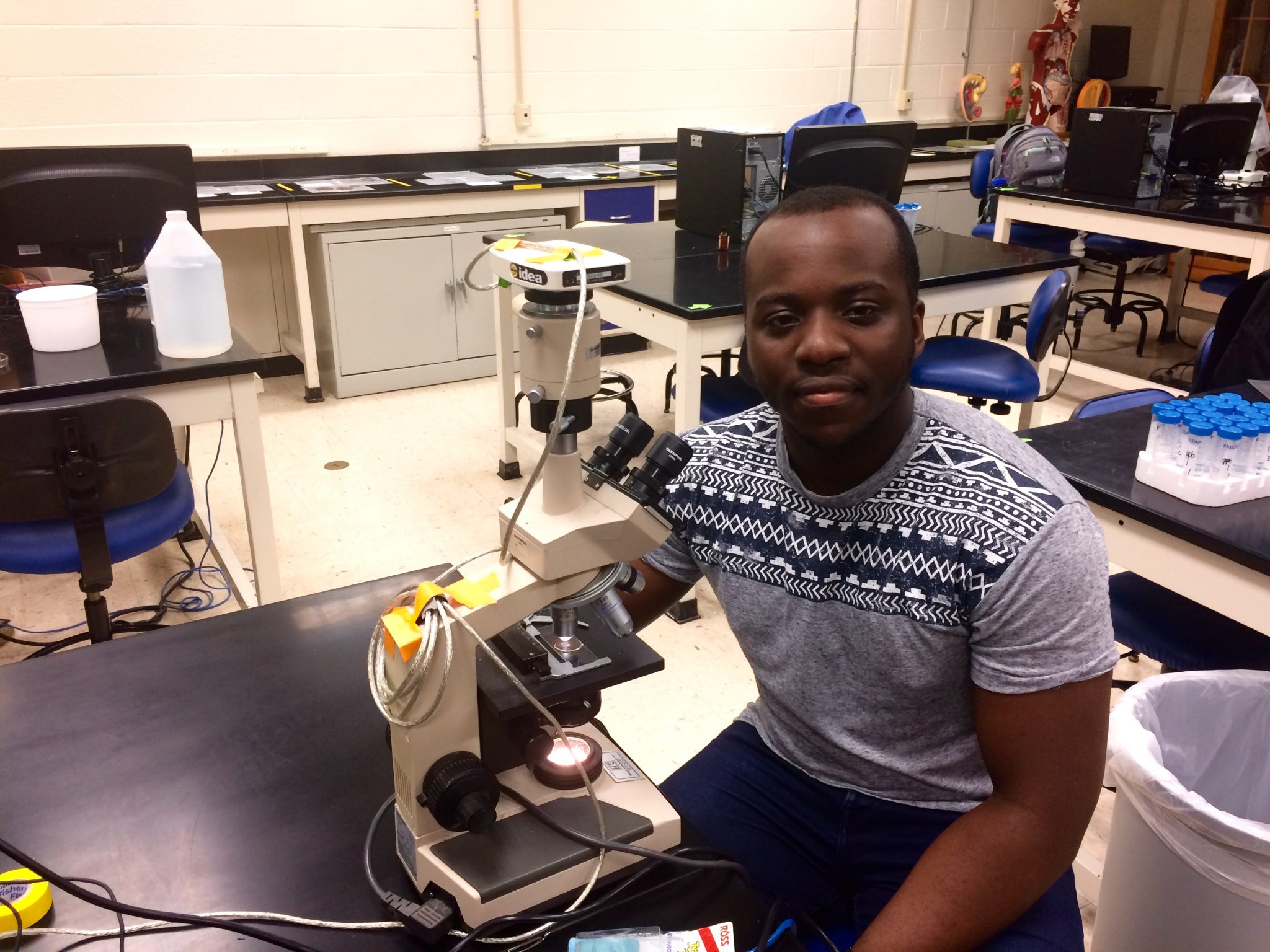 Eric Adjei-Danquah is sitting on a lab bench by a microscope. He is wearing a light grey shirt.