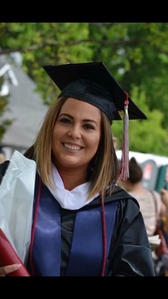 Nicole Enderica is wearing a graduation cap and gown in front of some green trees. She has a blue stole, and is carrying a white bag.