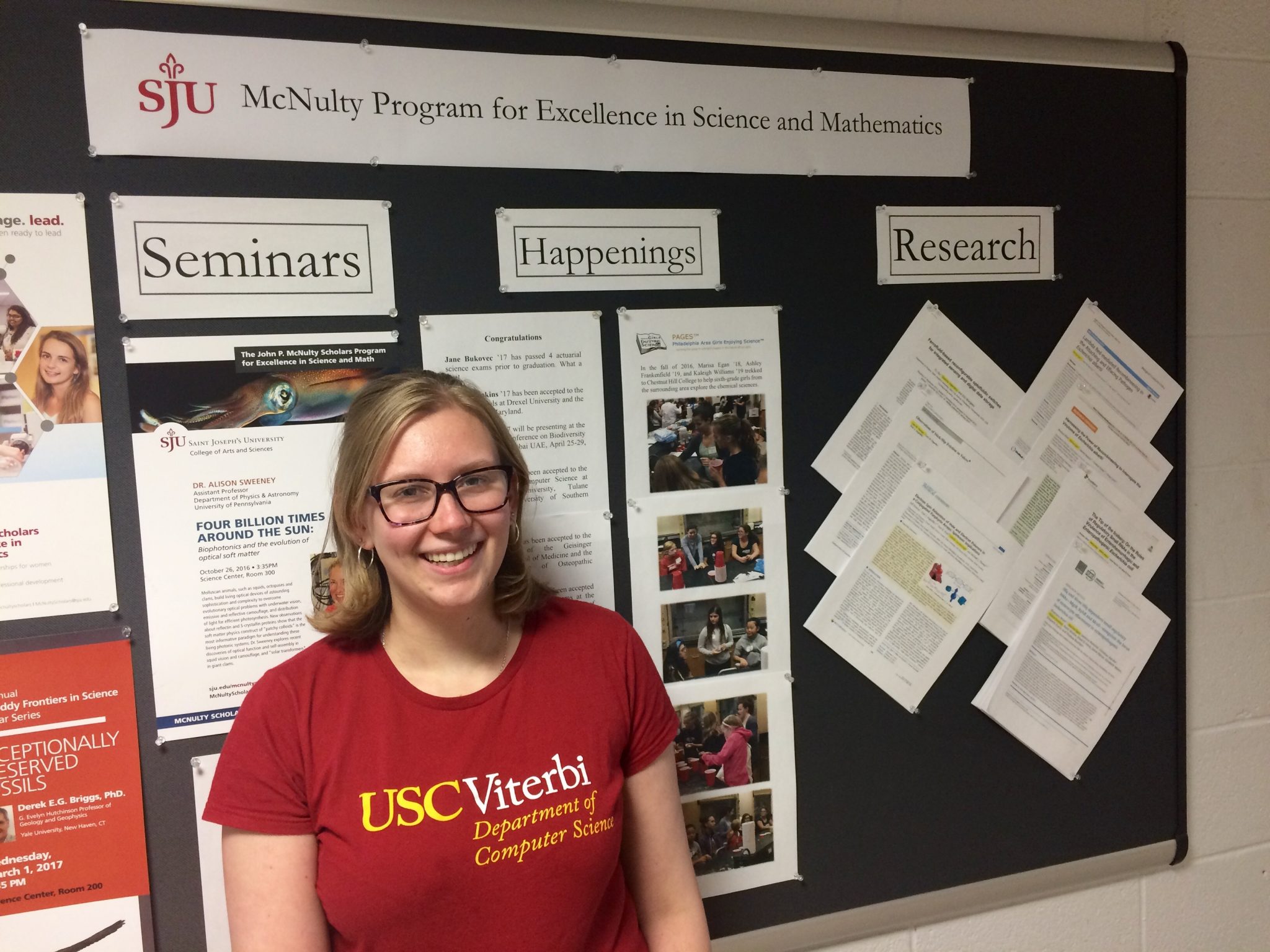 Sarah Cooney is smiling in front of a board reading, "SJU McNulty Program for Excellence in Science and Mathematics." She is wearing a red shirt and glasses.