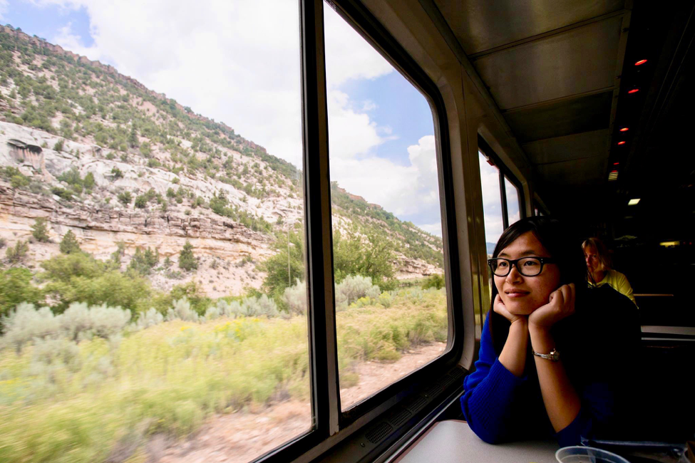 Wei Cai is staring out of the window of a train passing some vegetation. She is wearing a blue top and glasses.