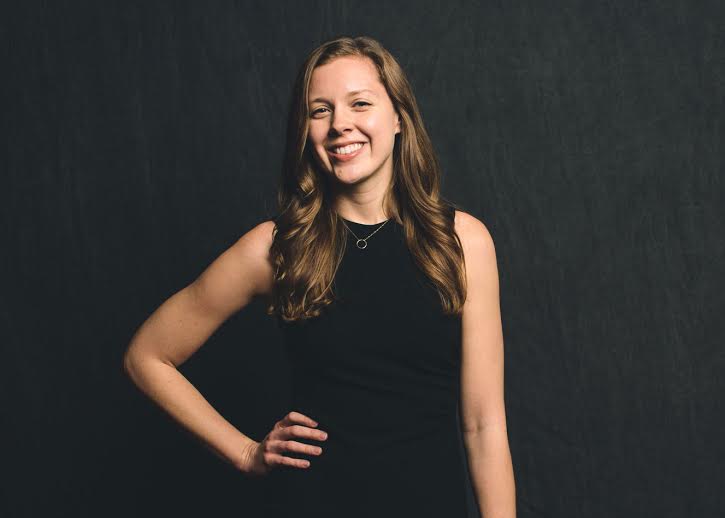 Suzanne Cotter is smiling with one hand on her hip in front of a black background. She is wearing a black dress.