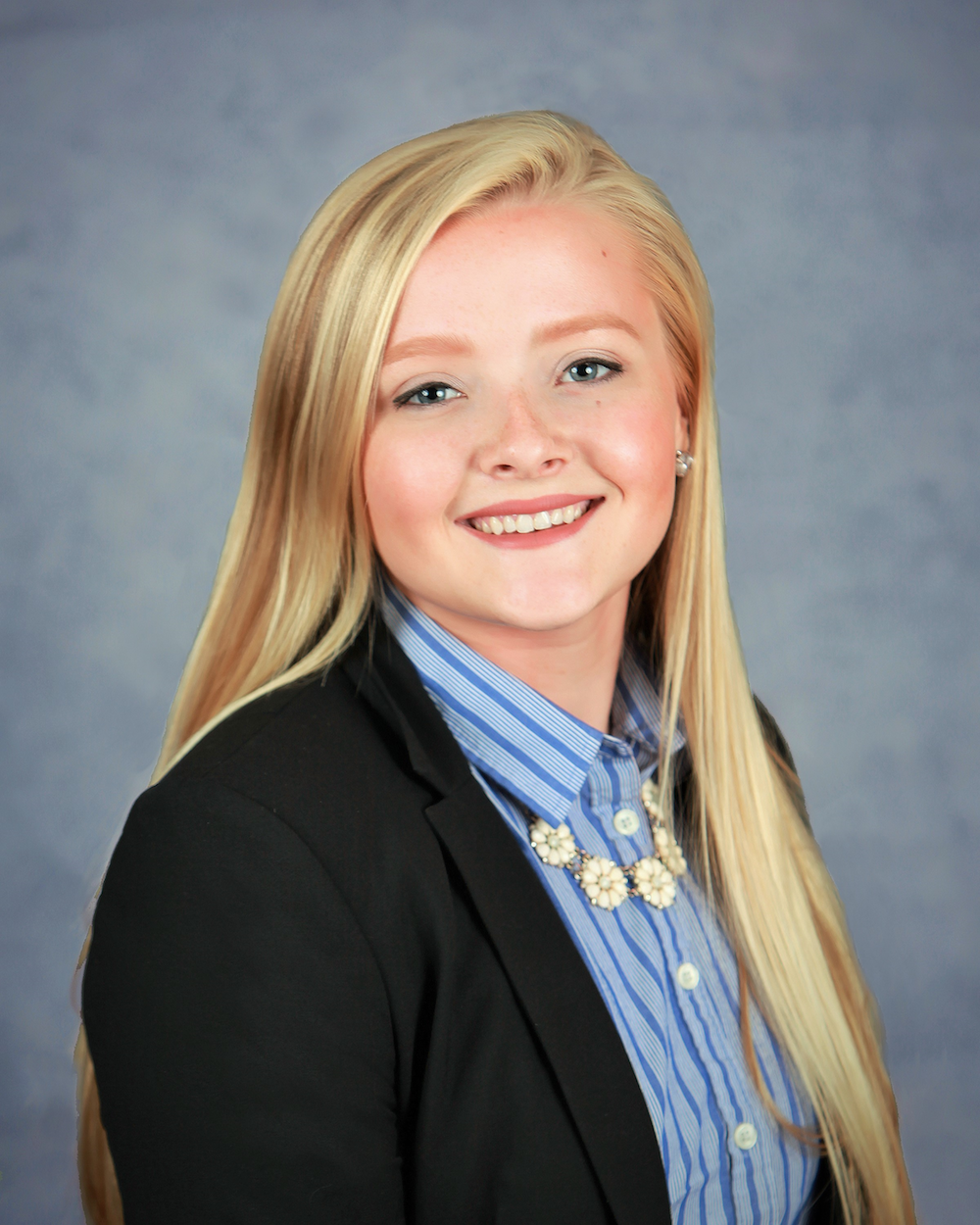 Cait English is smiling in front of a blue background. She is wearing a dark blazer with a blue, striped shirt, and a white floral necklace.