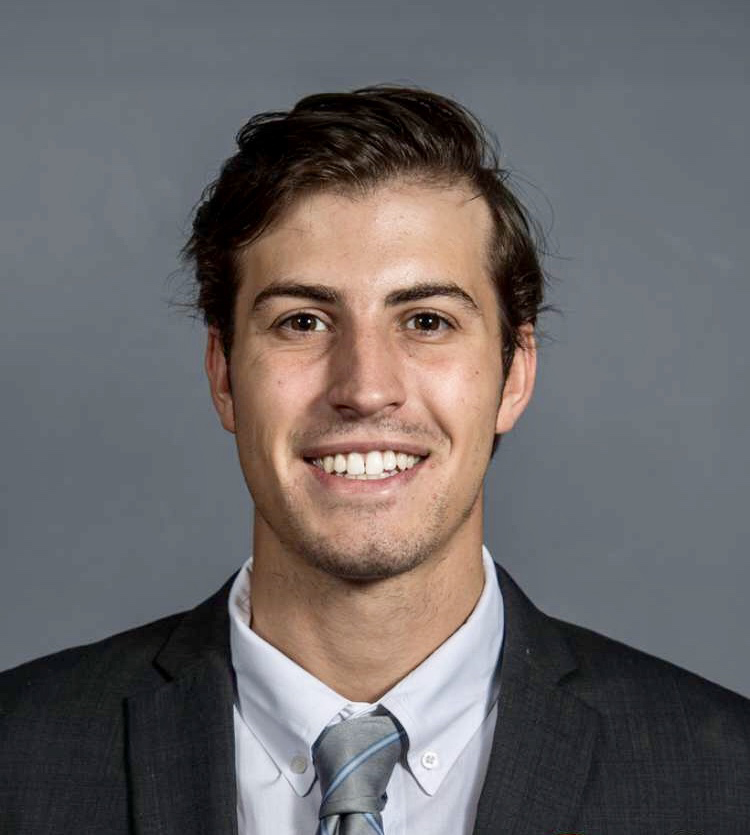 Harrison Zucco is smiling in front of a grey background. He is wearing a dark suit and a grey, striped tie.