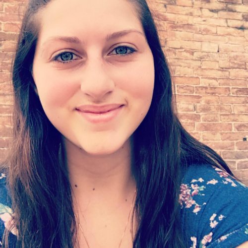 Alyssa LaMont is smiling in front of a brick wall. She is wearing her hair half up half down, and a floral top.