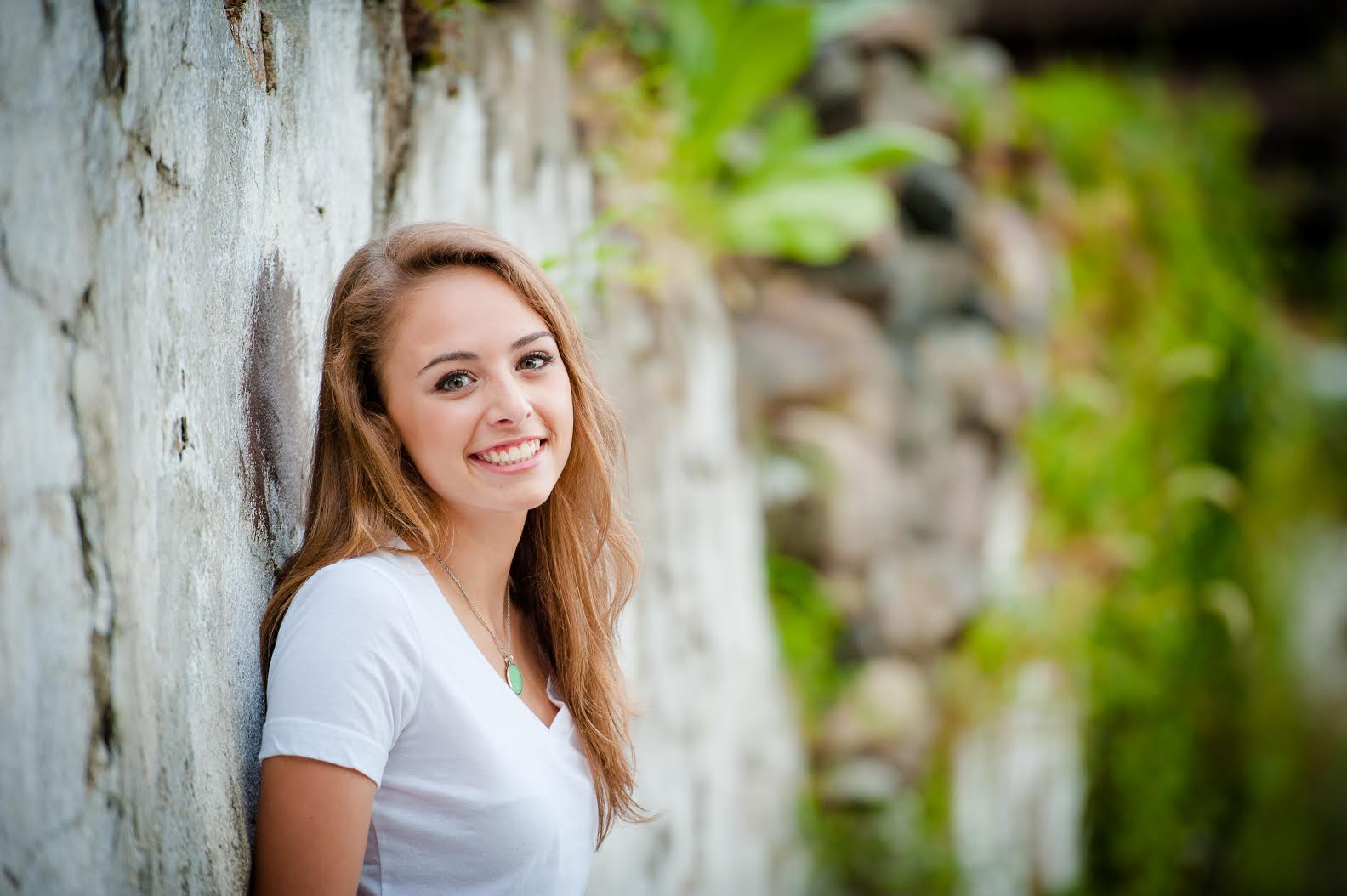 Paige Santiago is leaning against a stone wall in front of green, lush plants. She is wearing a white top and smiling broadly.