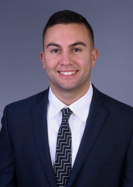 Colin McDonald is smiling in front of a grey background. He is wearing a dark suit with a tie with a zig-zag pattern.