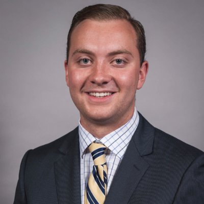 Michael Baldini is standing in front of a grey background. He is wearing a dark suit with a check shirt and a striped tie.