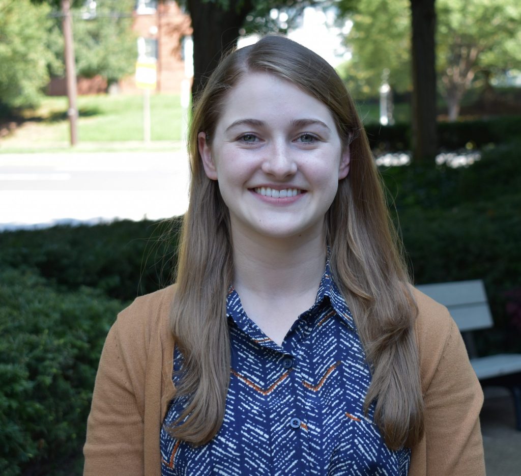 Emily McClellan is smiling in a park. She is wearing an orange cardigan and a blouse striped orange and blue.