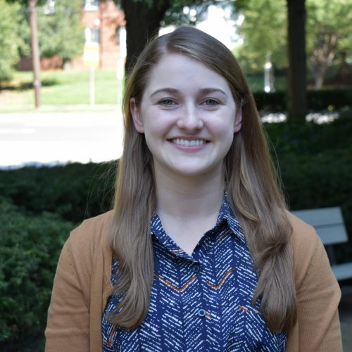 Emily McClellan is smiling in a park. She is wearing an orange cardigan and a blouse striped orange and blue.