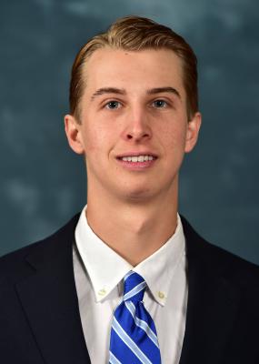 Austin Smith is smiling in front of a blue background. He is wearing a dark suit with a blue, striped tie.