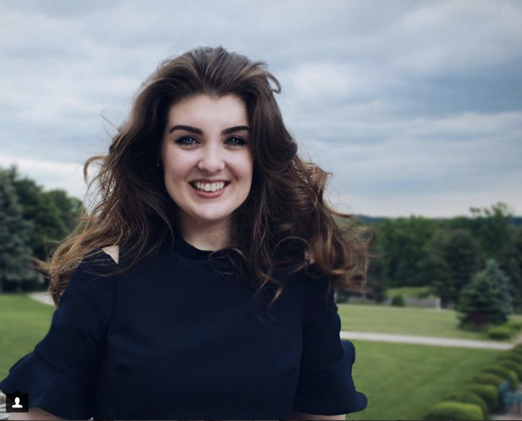 Caitlyn Sottile is smiling in front of a tree-lined background. She has very wavy hair, and is wearing a dark top.