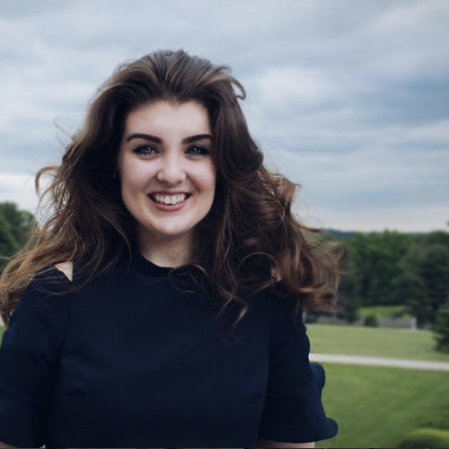 Caitlyn Sottile is smiling in front of a tree-lined background. She has very wavy hair, and is wearing a dark top.