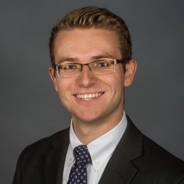 Jon Lemanski is smiling in front of a grey background. He is wearing a dark suit and square-patterned tie.