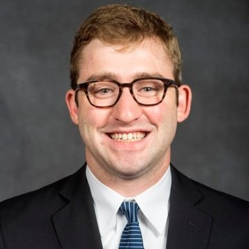 AJ Buono is smiling in front of a grey background. He is wearing glasses, a dark suit, and a striped tie.