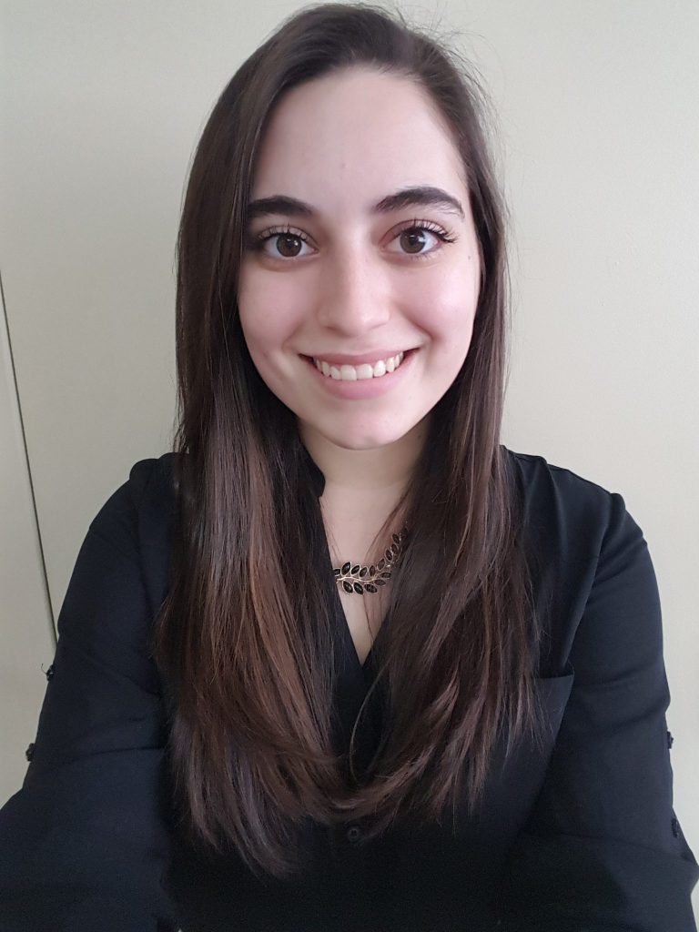 Juliana Ciccozzi is smiling in front of a white wall. She is wearing a dark top with a necklace, and has her hair straight.