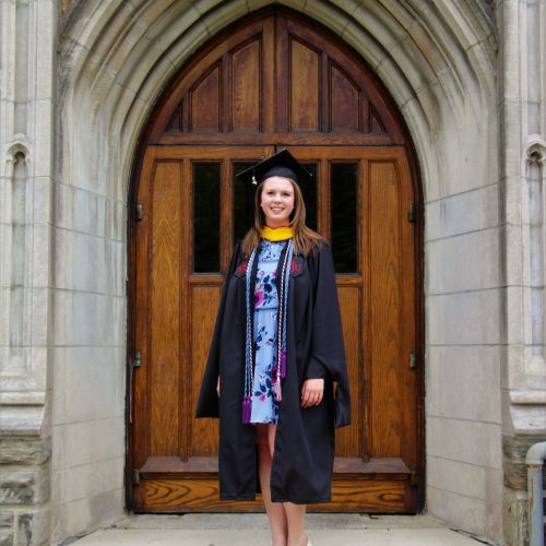 Daire Ryan is standing in front of an arched door from Barbelin. She is wearing a graduation cap and gown. She is wearing multiple honor cords, and a blue, floral dress.