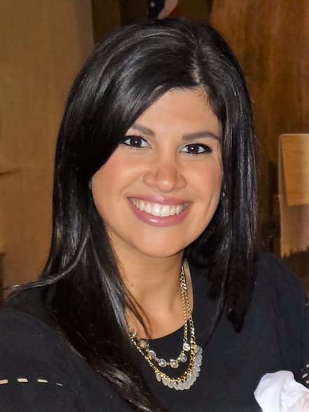 Giana Alongi is smiling in front of a brown room. She is wearing a dark top and gold necklaces.