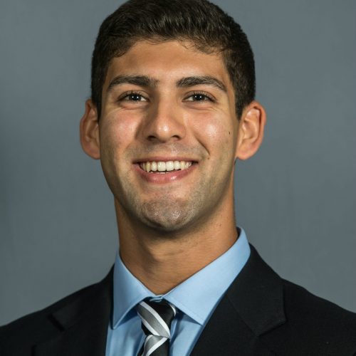 Anthony Savarese is smiling in front of a blue background. He is wearing a dark suit with a pale blue shirt and a striped tie.