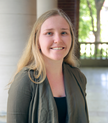 Kaitlyn Martin is smiling in front of a background with white columns and blurred trees. She is wearing a dark top and a green cardigan.