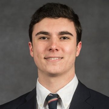 Brendan Garry is smiling in front of a grey background. He is wearing a suit with a striped tie.