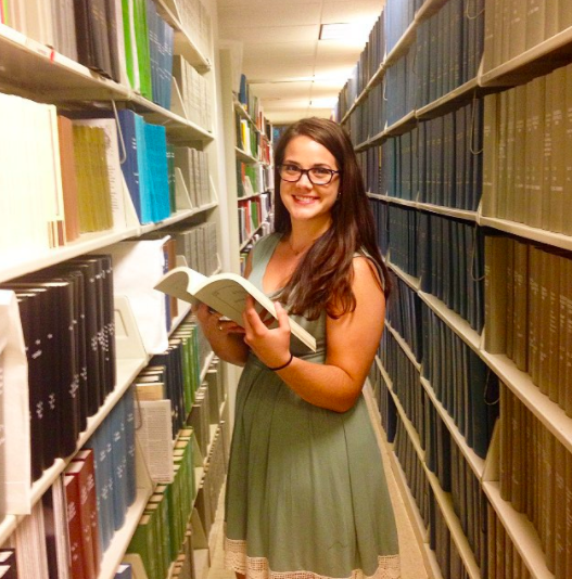 Katherine Grygo is standing between two library shelves, with a book opened in her hands. She is wearing a green dress and glasses.