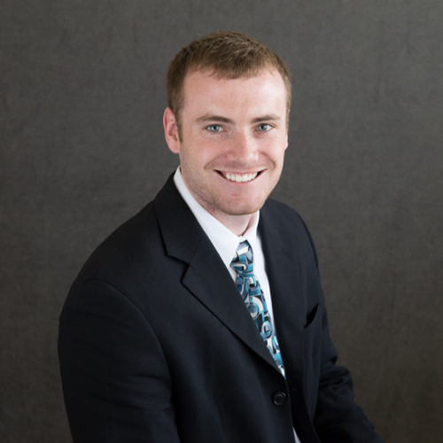 Corey MacDonald is facing the right, smiling in front of a dark background. He is wearing a dark suit with a patterned, teal tie.