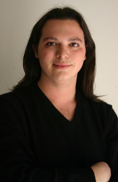 Gregory Day is smiling in front of a white background. He has dark dark hair, and is wearing a dark top.