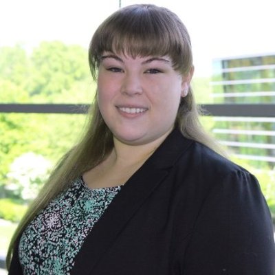 Lindsay Miller is smiling in front of a blurred background of trees and an office building. She is wearing a dark blazer with a patterned top.