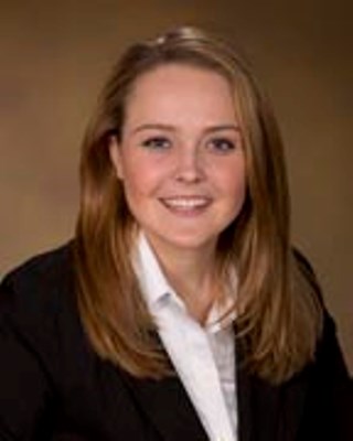 Kristen Rupp is smiling in front of a brown background. She is wearing a dark blazer and a white shirt.