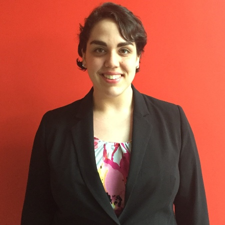 Sara Serpe is smiling in front of a red background. She is wearing a black blazer with a floral top, and her hair is wavy and short.