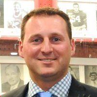 Keith Griesbaum is smiling in front of football photos. He is wearing a check shirt with a dark blazer and pale blue tie.