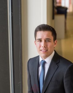 John Verzi is a young man sitting by a Haub window. He is wearing a dark suit with a blue tie.