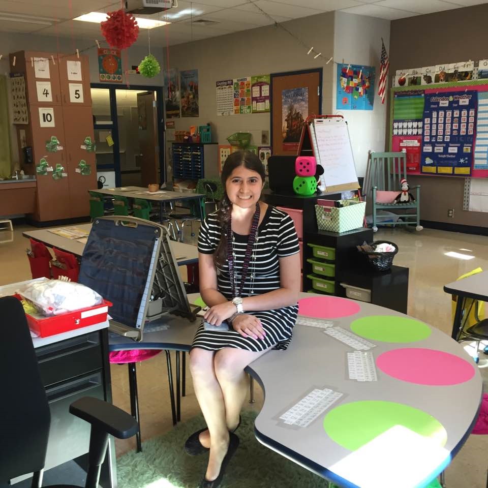 Melanie Solano is sitting on a curved table in an elementary school classroom. Her hair is in braids, and she is wearing a striped dress.